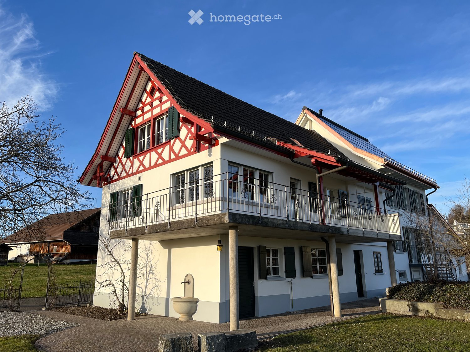 Two-story house with white walls, red trim, and a balcony on the upper level. The house features multiple windows with shutters and a small garden area in front.