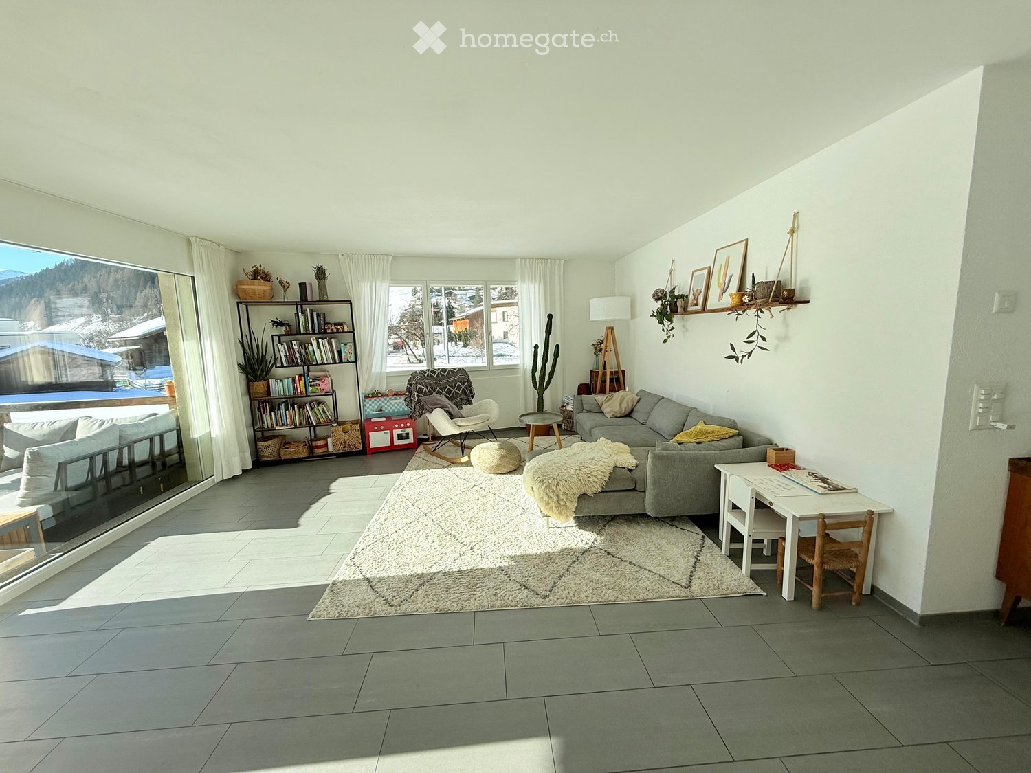 Bright living room with large windows offering a scenic view, featuring a grey sectional sofa, a white armchair, a small round table, a bookshelf, a floor lamp, and a white shaggy rug.
