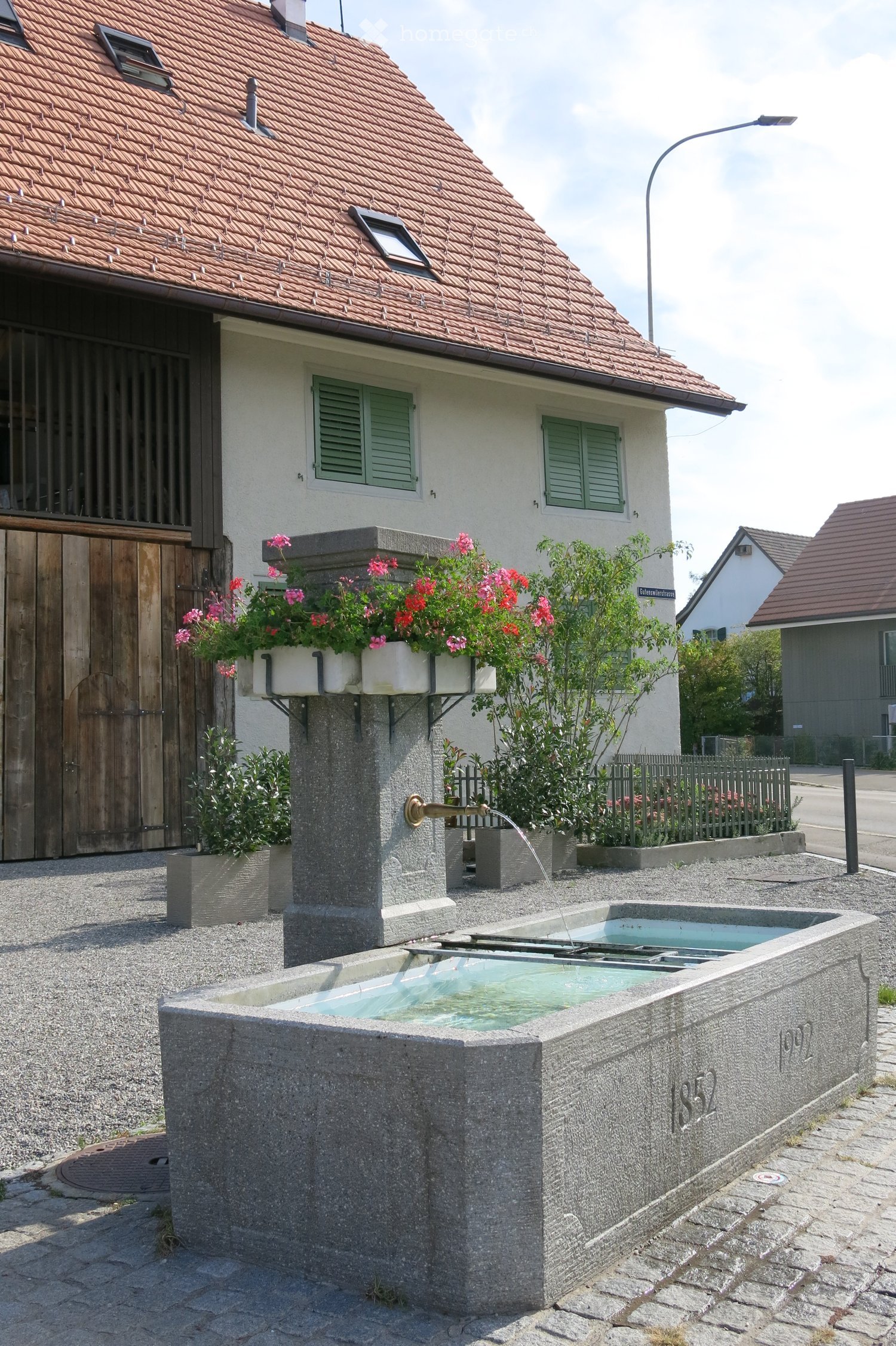 A stone fountain with flowing water, surrounded by greenery and flowers, with a house in the background featuring a wooden garage and green shutters.