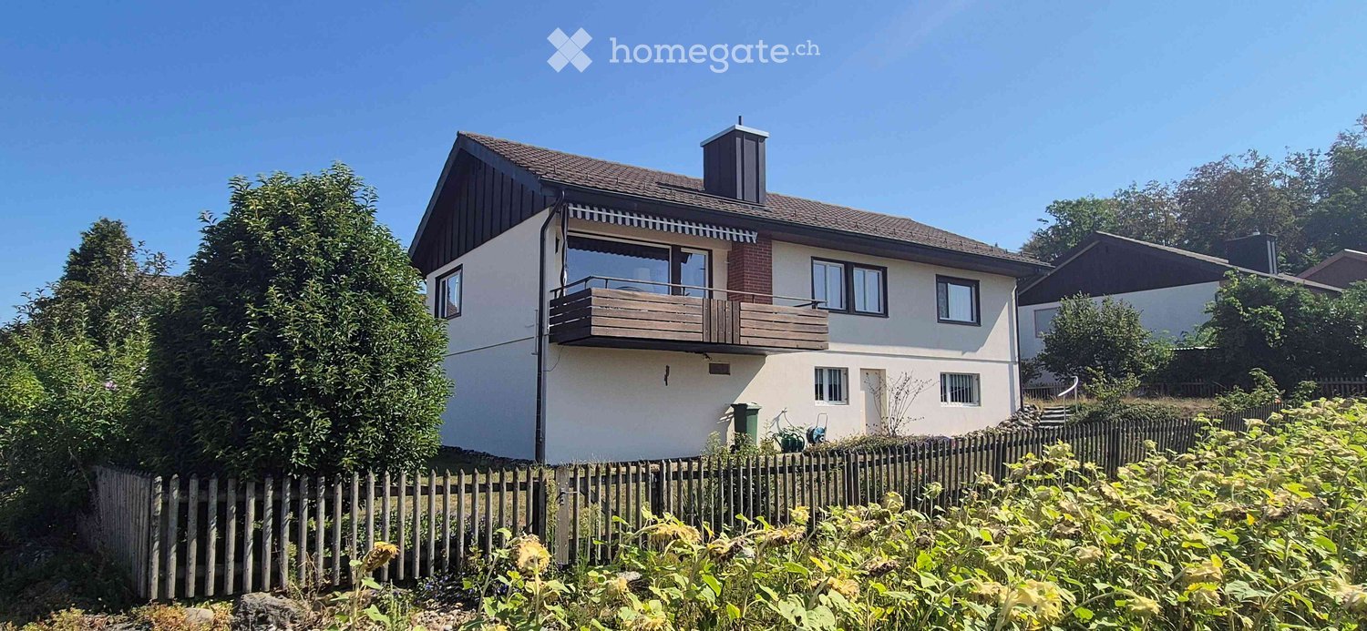 Two-story house with a balcony, surrounded by greenery and a wooden fence.