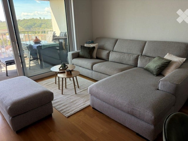 Modern living room with grey sectional sofa, ottoman, coffee table, side table, and a view through large windows.