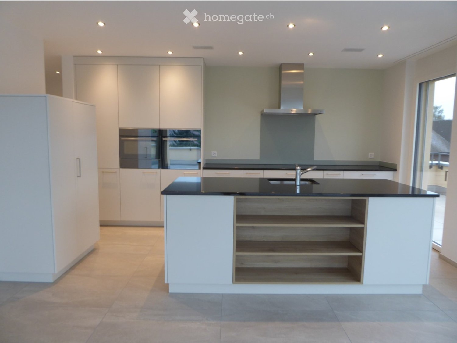 Modern kitchen with white cabinets, black countertops, and a central island with open shelving. Features include a stainless steel range hood, built-in oven, and a sink with a modern faucet.