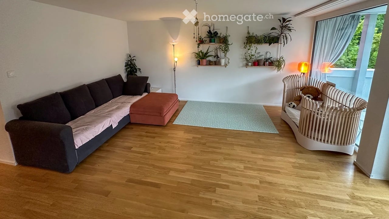 Living room with wooden flooring, a dark brown sectional sofa with pink throw, an orange ottoman, a white baby crib with stuffed animals, floor lamp, table lamp, and wall-mounted shelves with plants.