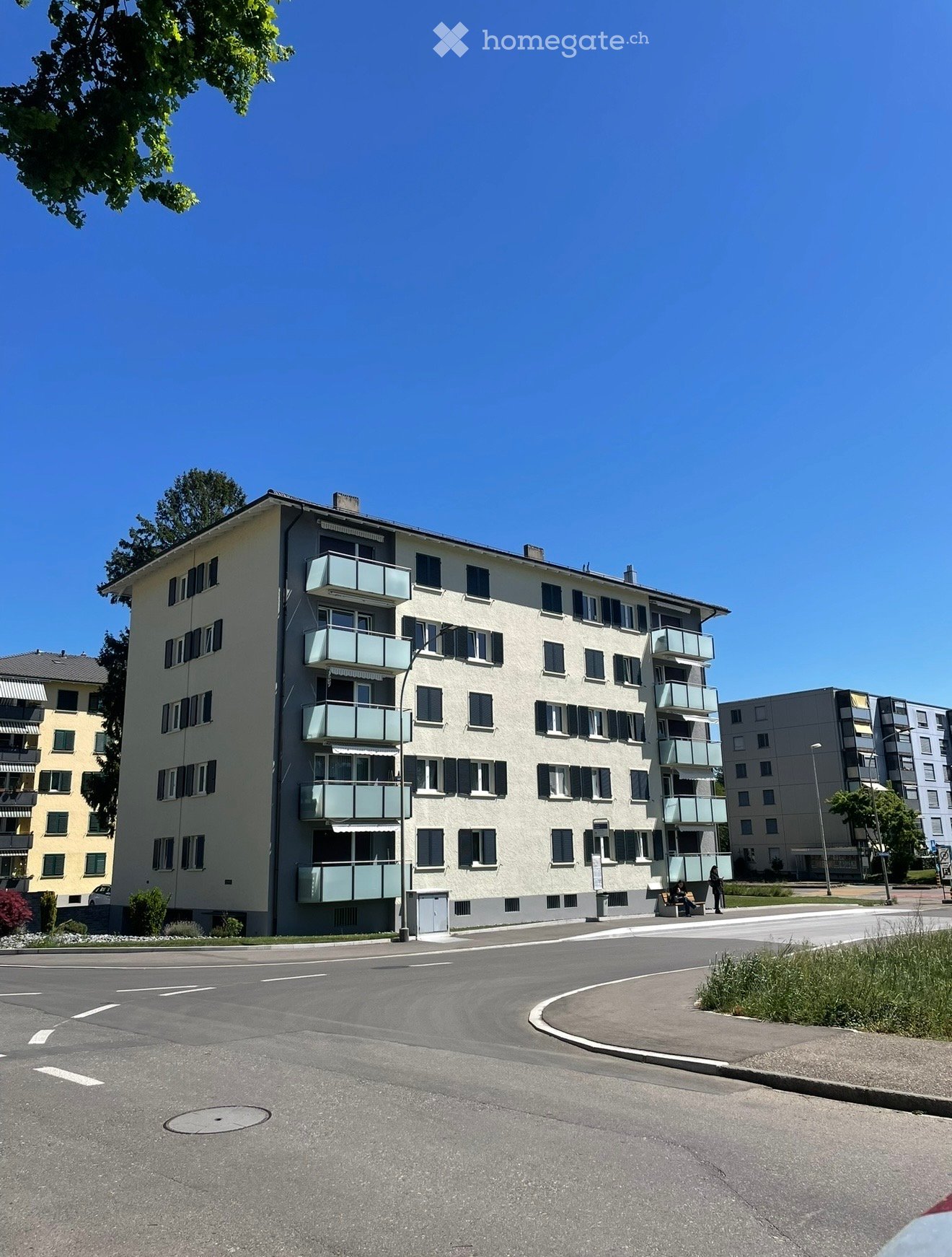 Multi-story residential building with balconies, windows, and shutters, surrounded by a paved area and greenery.