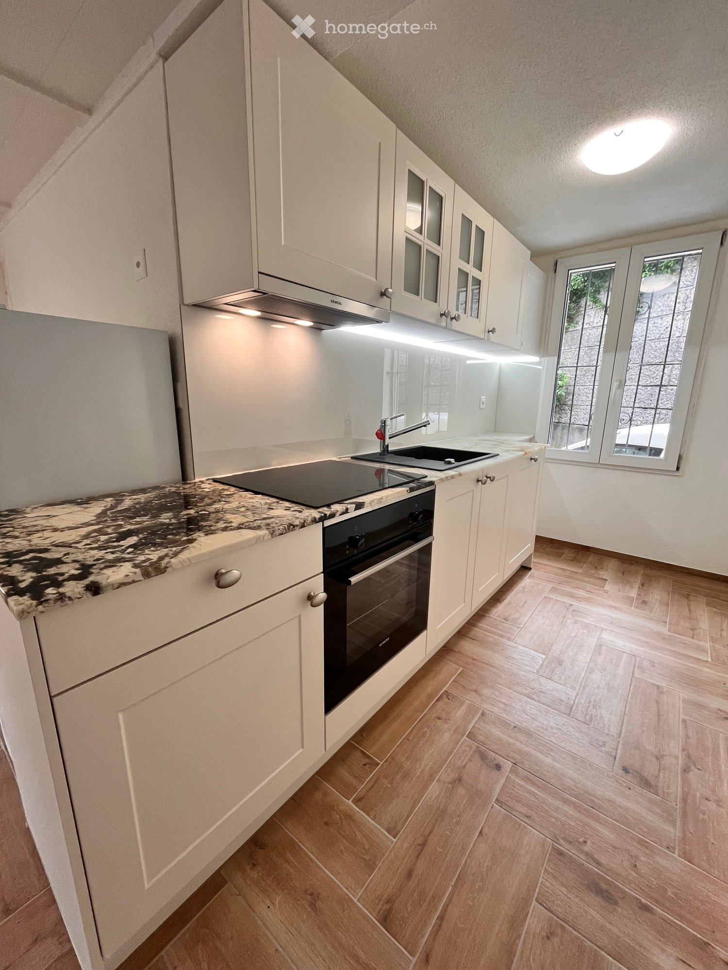Modern kitchen with white cabinets, marble countertop, built-in oven, sink, and range hood. Wooden flooring and large windows provide natural light.