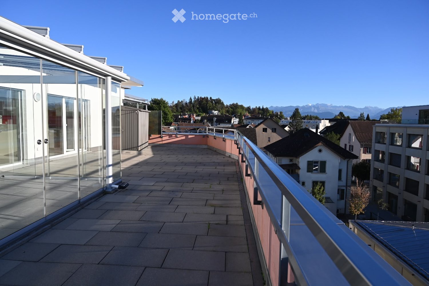 Modern balcony with tiled flooring, glass railings, and a view of surrounding buildings and mountains.