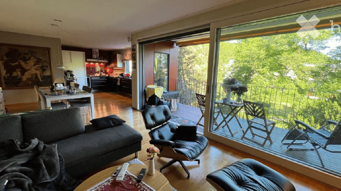 Modern living room with large windows and a balcony overlooking greenery. Features a grey sofa, coffee table with remote controls, leather chair, and a small table with a vase of flowers. The adjacent kitchen area is visible with a dining table and chairs