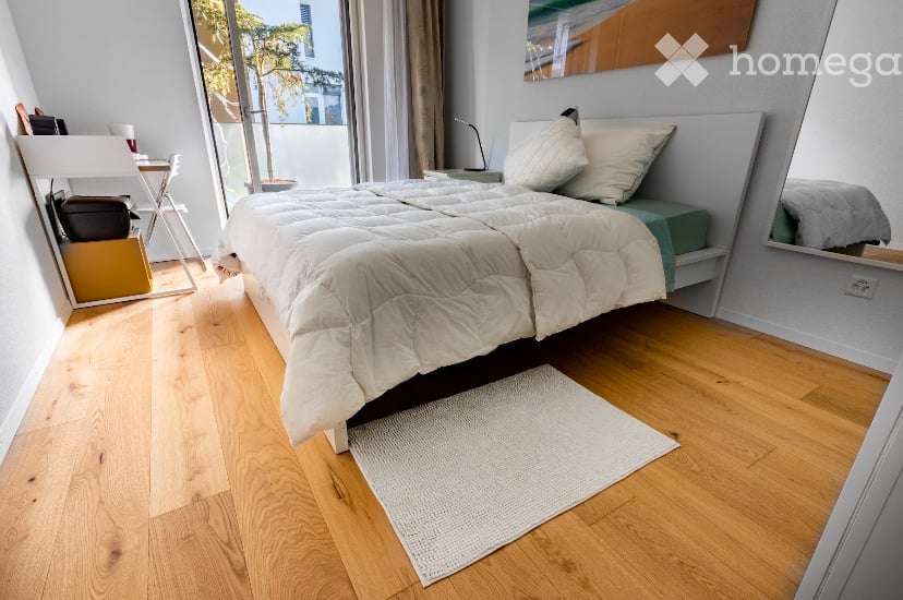 Cozy bedroom with wooden flooring, a large bed with white bedding and pillows, a nightstand with a lamp, a mirror, a small chair, and a table with items on it.
