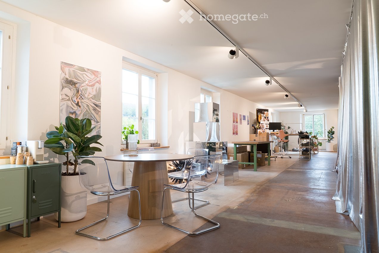 Bright, open room with large windows, a round wooden table with four modern chairs, a green cabinet, and several potted plants. The space includes a workspace area with a desk, computer, and additional seating.