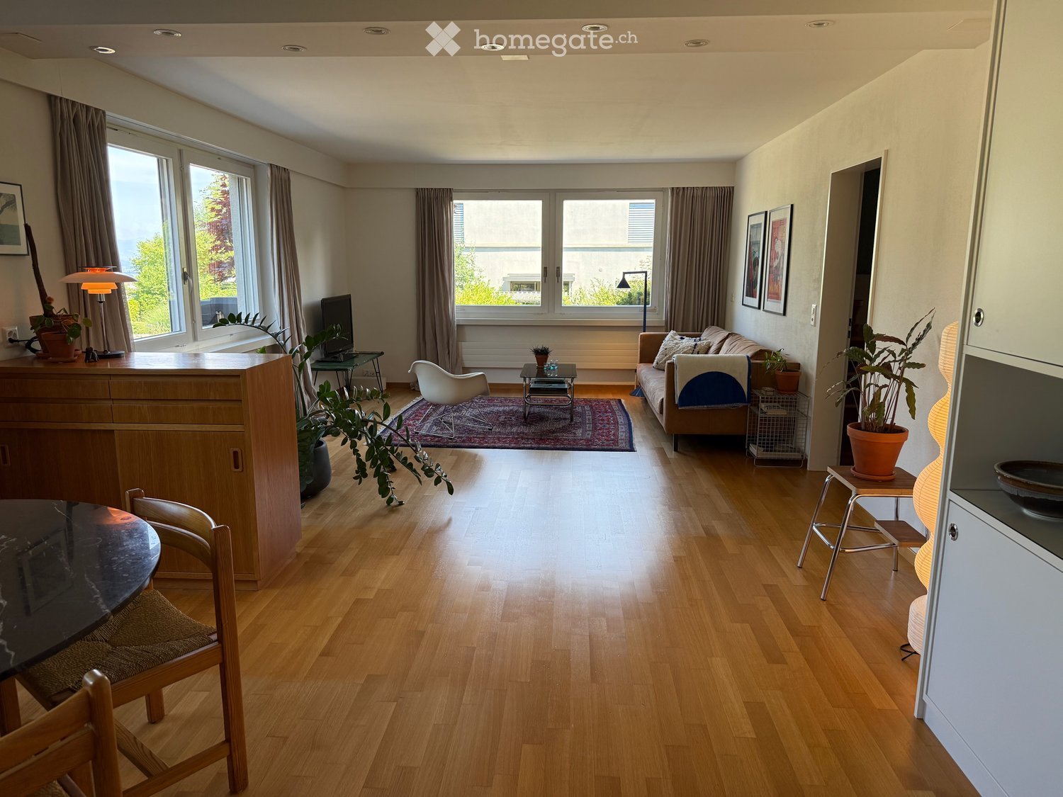 Bright living room with wooden flooring, large windows, and modern furniture including a sofa, coffee table, and chairs.