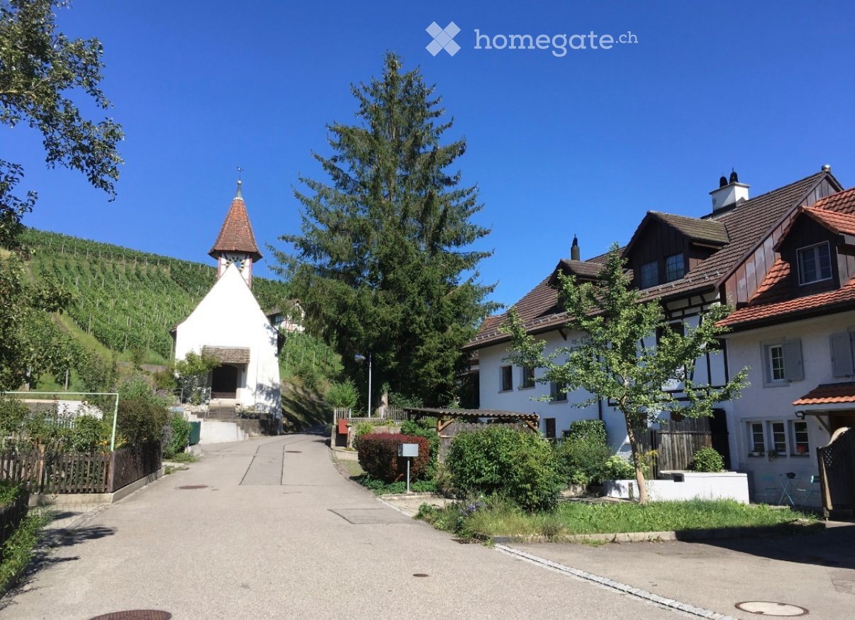 A small village scene with a white church featuring a pointed spire, surrounded by traditional houses with pitched roofs and wooden facades. The area is lush with greenery and trees, and a paved road leads through the village.