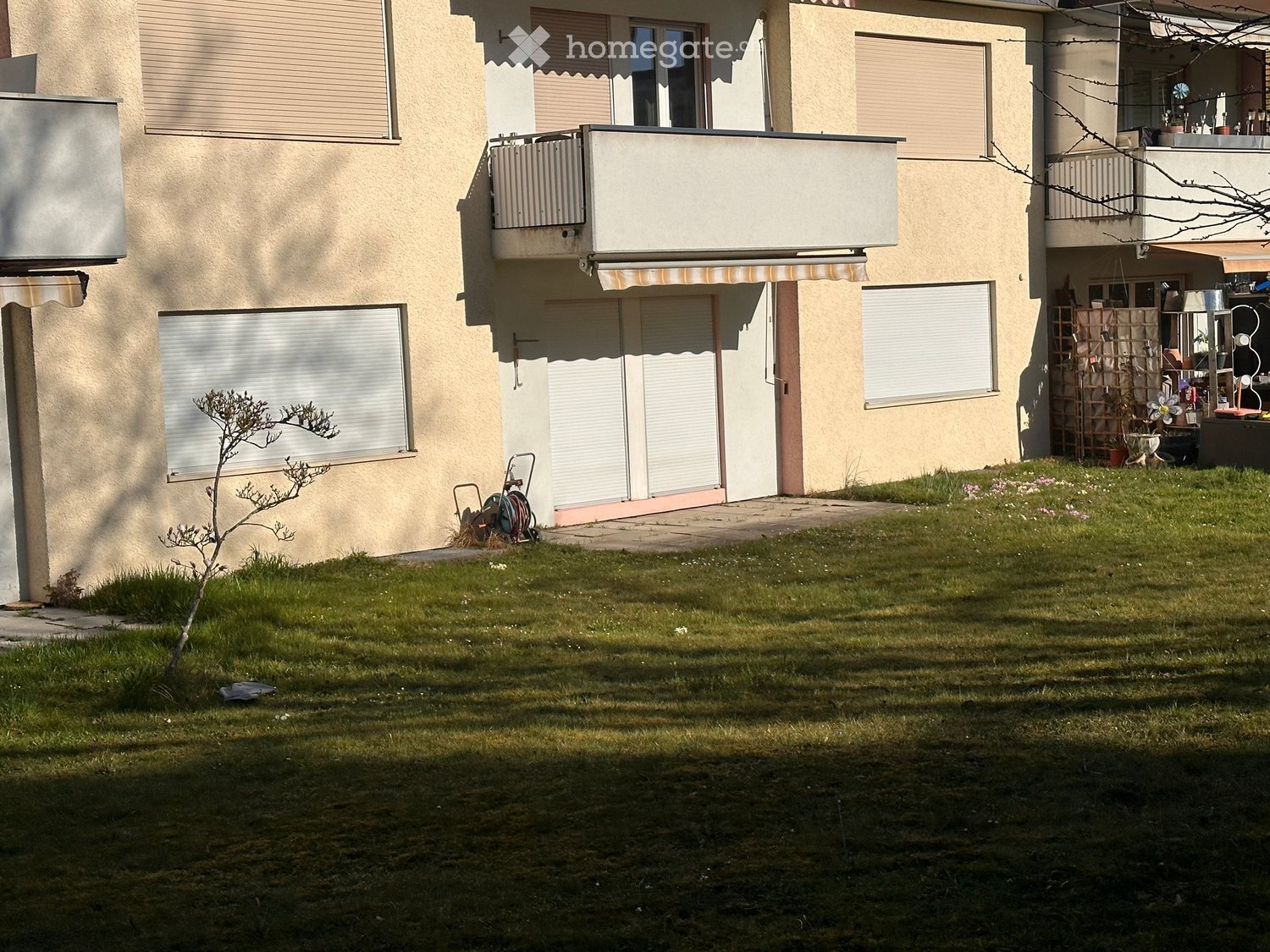 Front yard with grass, small tree, and a multi-story building with balconies and shutters in the background.