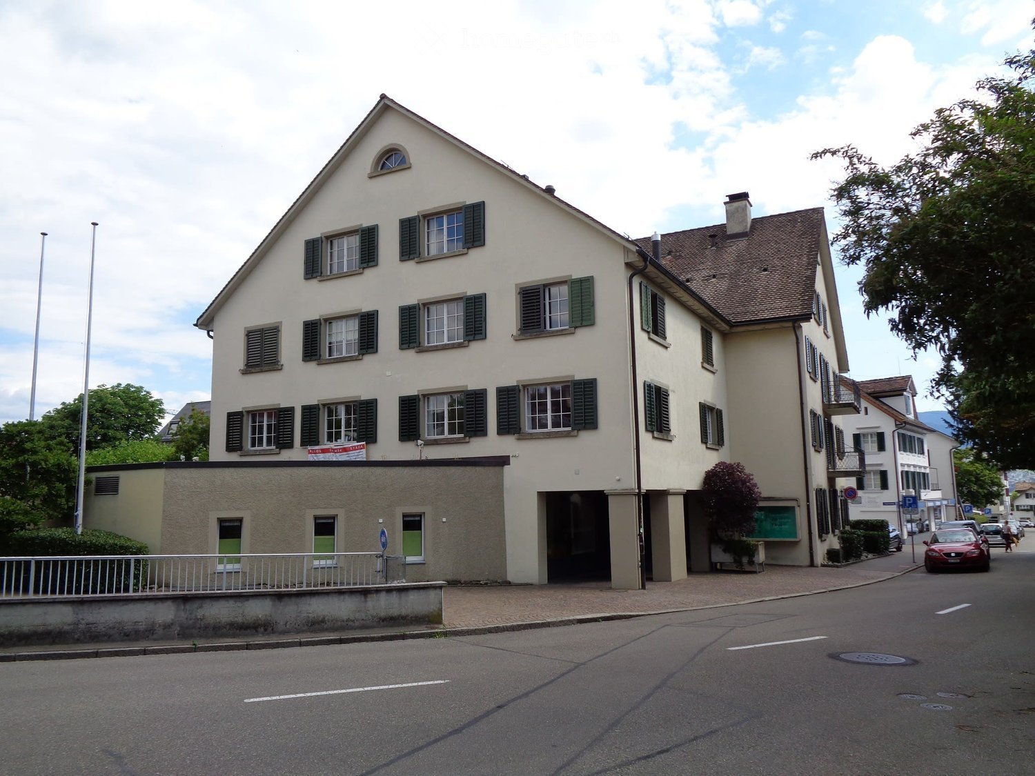 Two-story residential building with white facade, green shutters, and a garage entrance. The building has multiple windows and a pitched roof.