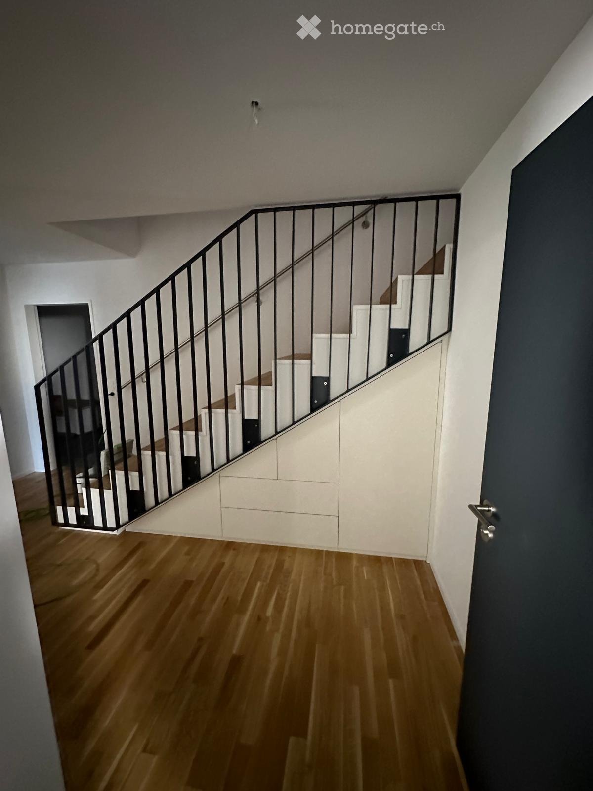 Modern hallway with wooden flooring, white walls, and a staircase with a black metal railing leading to an upper level.