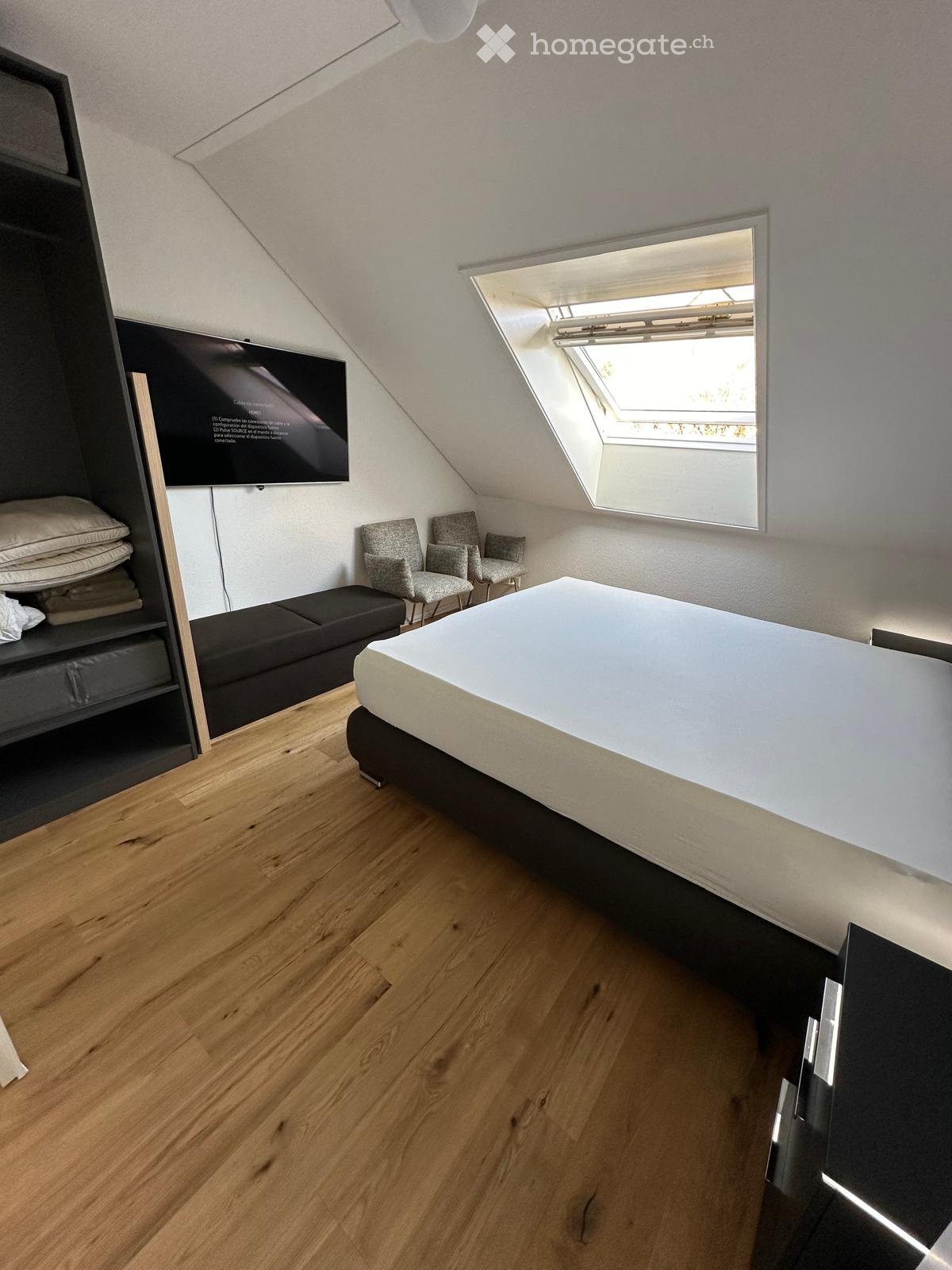 Modern attic bedroom with wooden flooring, a large bed, two chairs, a bench, and a built-in wardrobe with a TV screen.