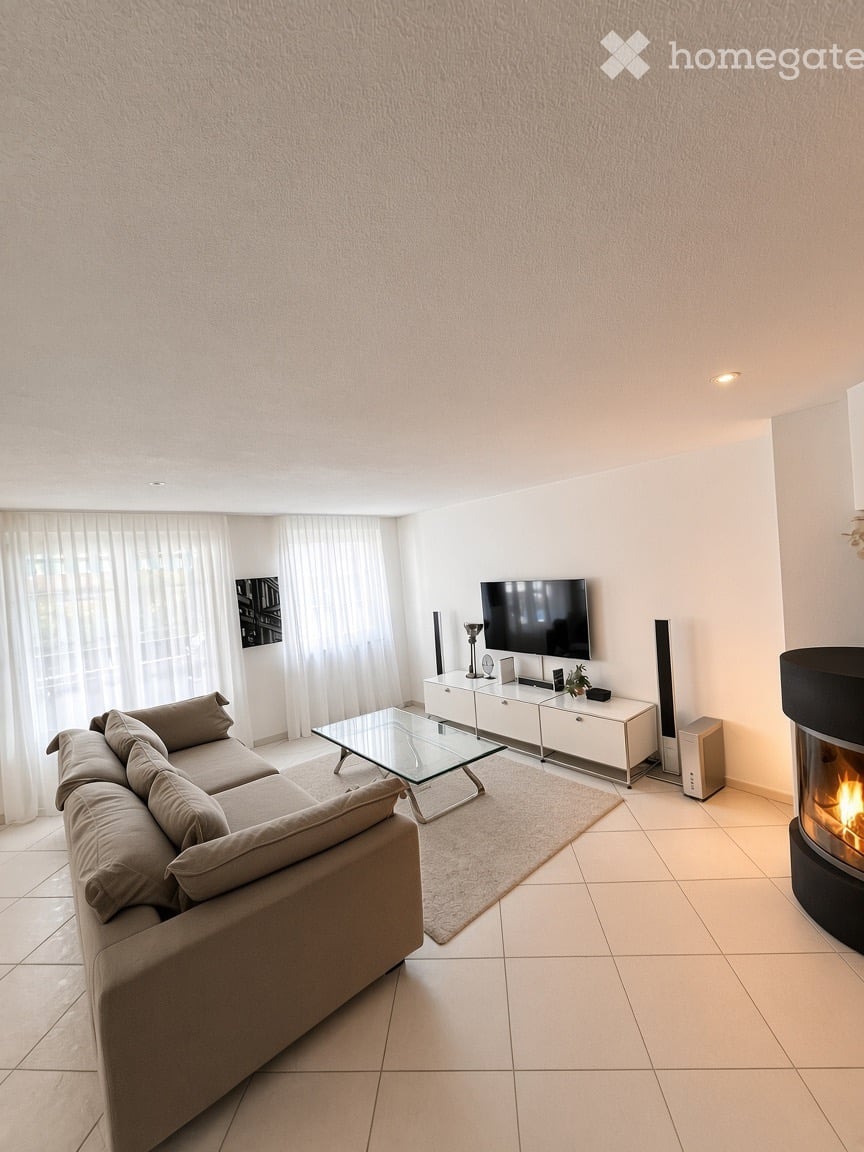Modern living room with a beige sofa, glass coffee table, flat-screen TV on a white cabinet, and a contemporary fireplace. The space features large windows with white curtains and a tiled floor.