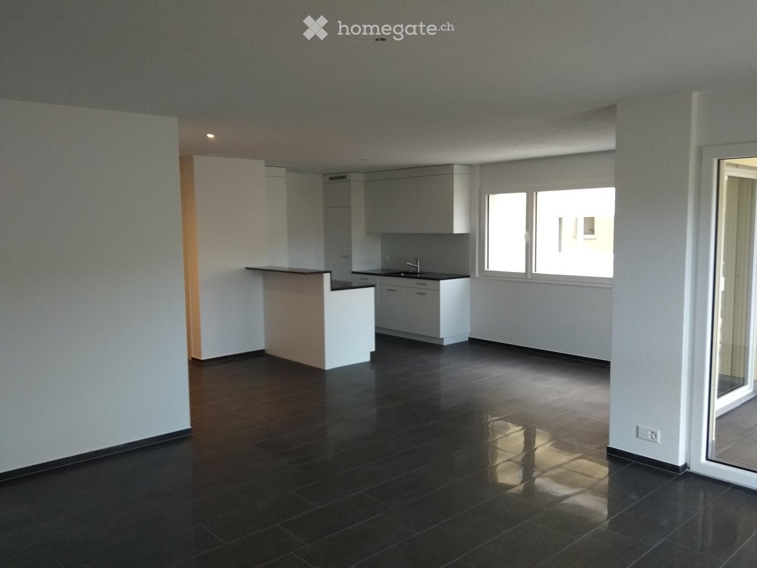 Empty room with dark tiled floor, white walls, and a small kitchen area with cabinets and a sink.