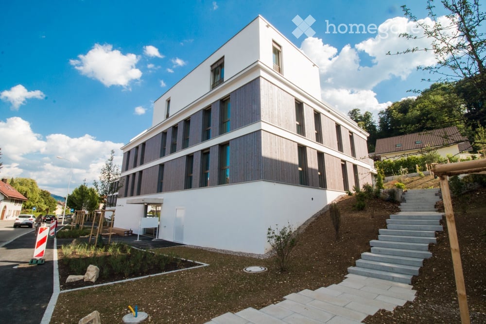 Modern two-story building with white and grey facade, large windows, raised ground floor, and landscaped surroundings.