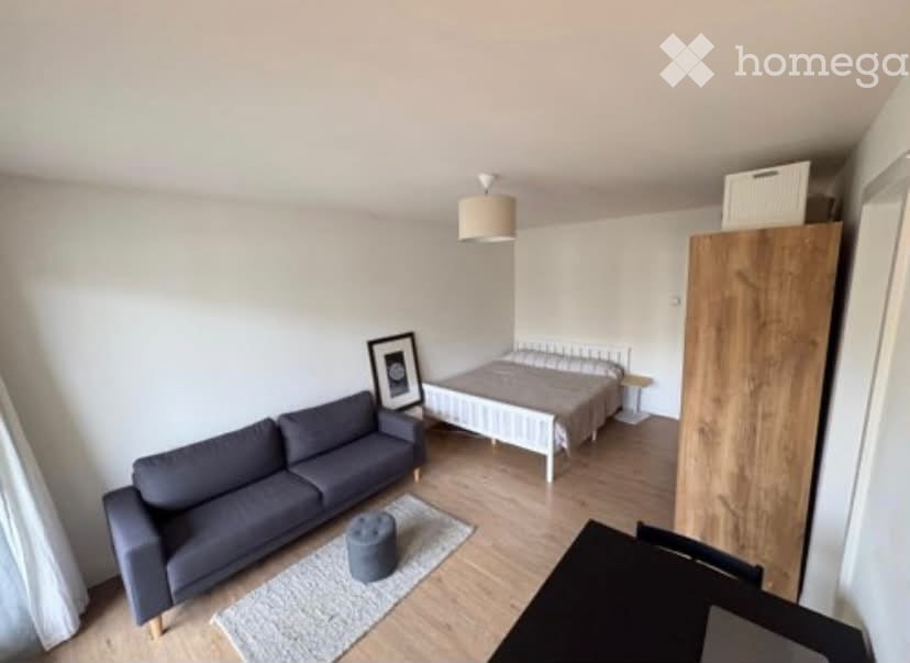 A modern, compact living space with a sofa, a small bed, a wardrobe, a picture frame, and a small round stool. The room features light wooden flooring and white walls.