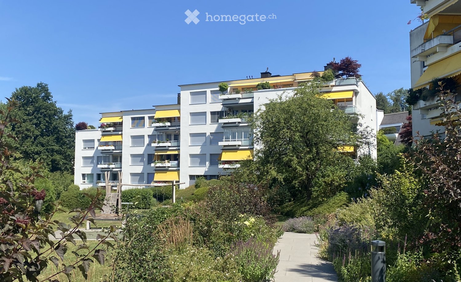 Modern multi-story residential building with white facades, yellow awnings, and balconies, surrounded by greenery and a landscaped garden area.