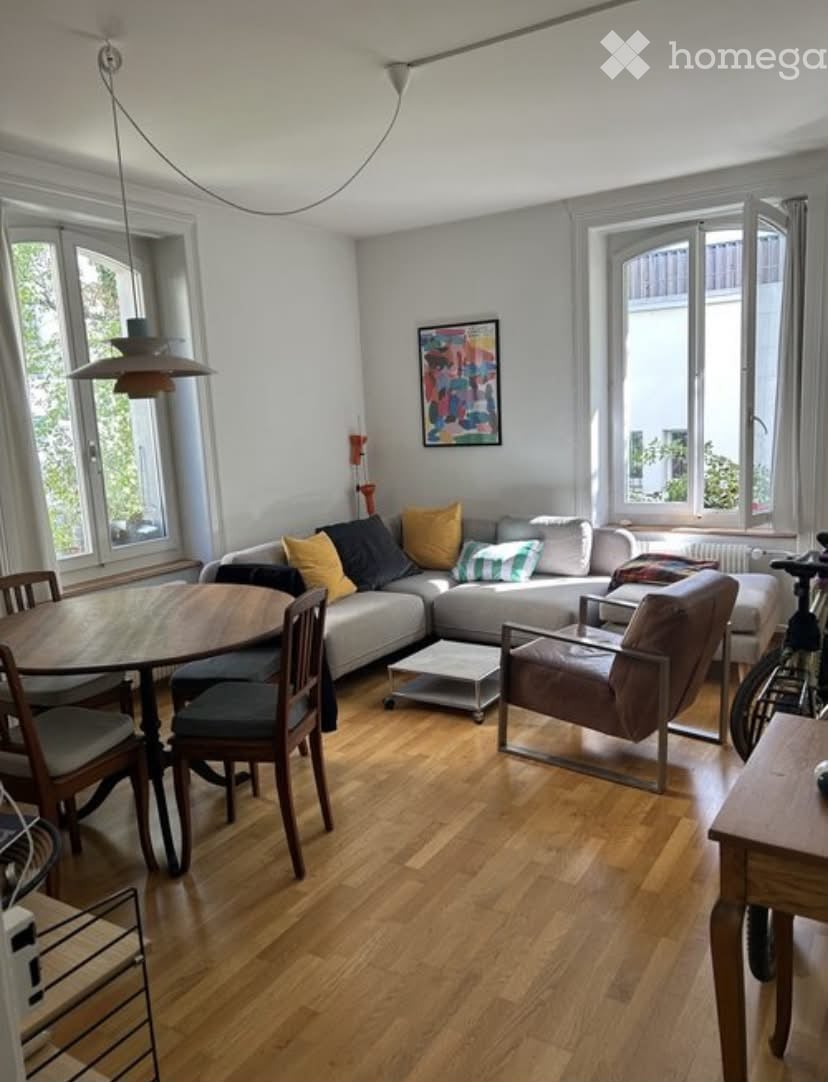 Bright living room with light grey sectional sofa, colorful throw pillows, wooden dining table with chairs, pendant light, and artwork on the wall.