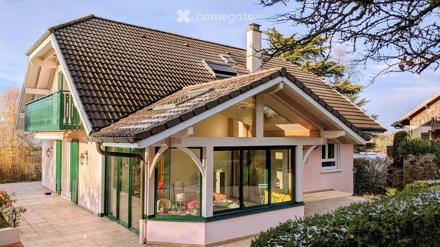 A modern two-story house with a tiled roof, green balcony, and large glass windows. The house features a pink exterior and is surrounded by a well-maintained garden with bushes and trees.