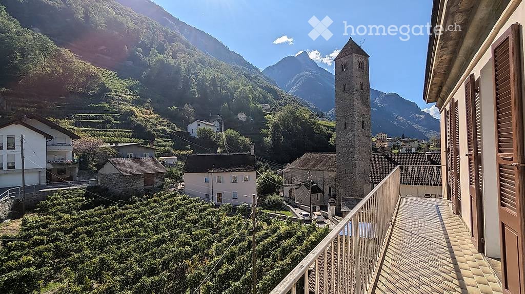 Balcony overlooking a picturesque village with stone houses, a tall tower, and lush green vineyards, surrounded by mountains.