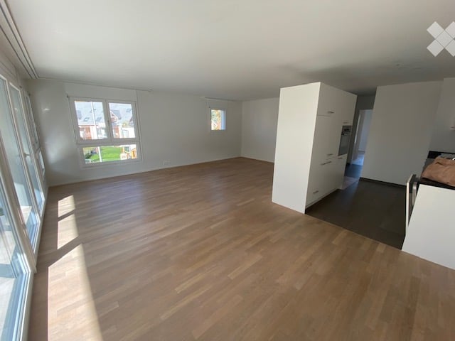 Empty room with wooden flooring, white walls, and large windows allowing natural light. The room is unfurnished and appears to be part of a modern interior design.
