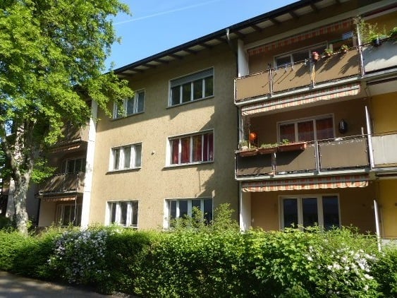 Multi-story residential building with balconies, large windows, and surrounding greenery.