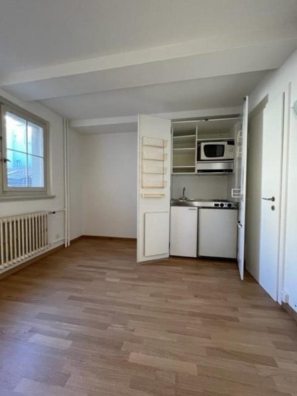 Empty room with wooden flooring, white walls, and a built-in kitchen area featuring cabinets, a microwave, and a sink.