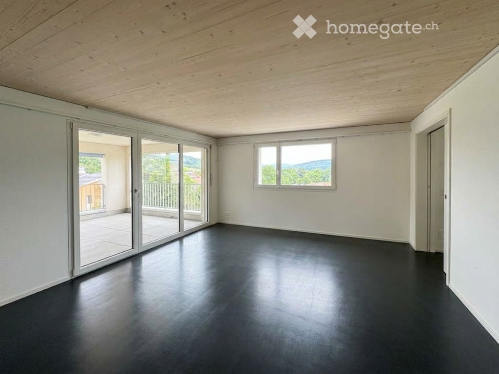 Empty room with white walls, dark flooring, large sliding glass doors leading to a balcony with a scenic view, and a window with a view of greenery and mountains.