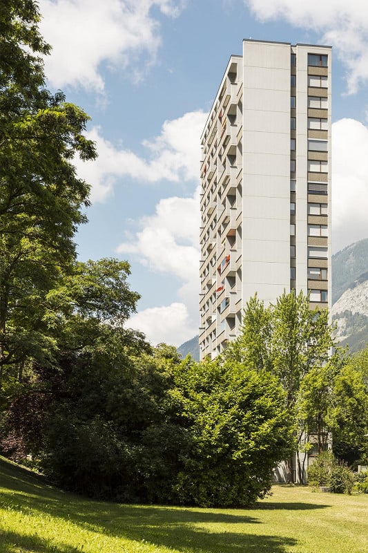 Modern high-rise apartment building with balconies, surrounded by lush green trees and a grassy lawn under a partly cloudy sky.