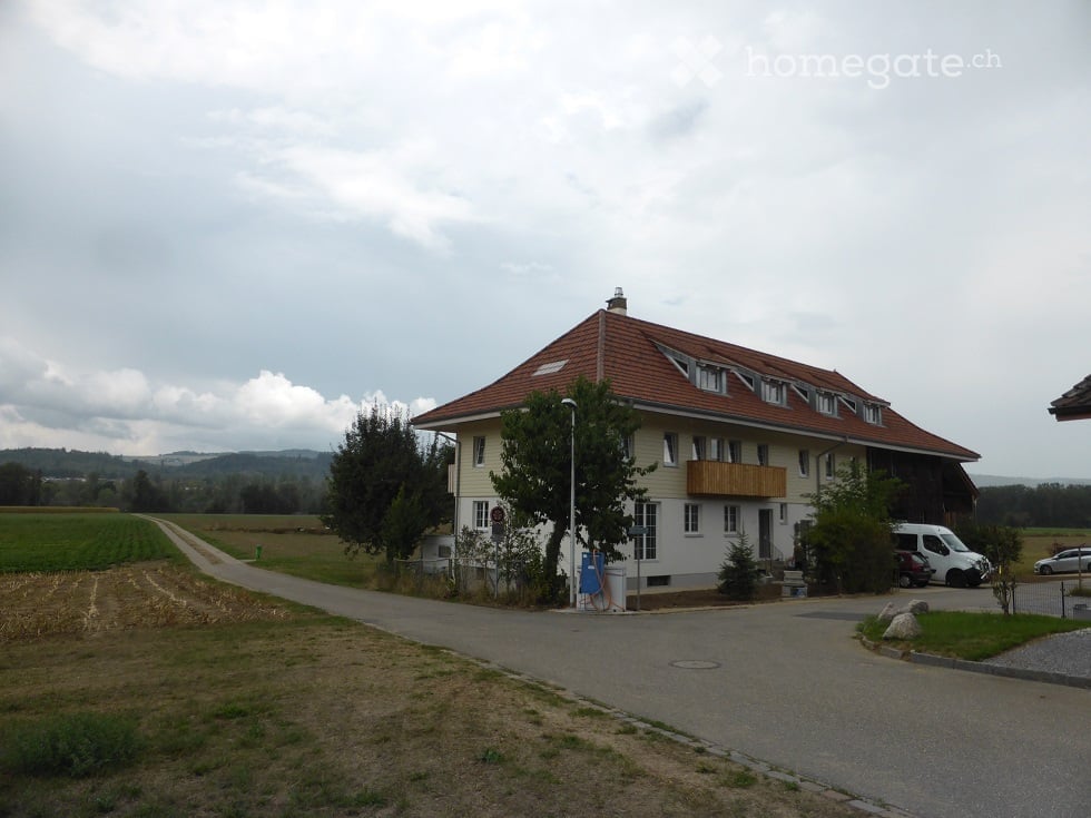 Two-story house with a red-tiled roof, white walls, and multiple windows. Features a balcony on the upper floor and a driveway with parked vehicles.