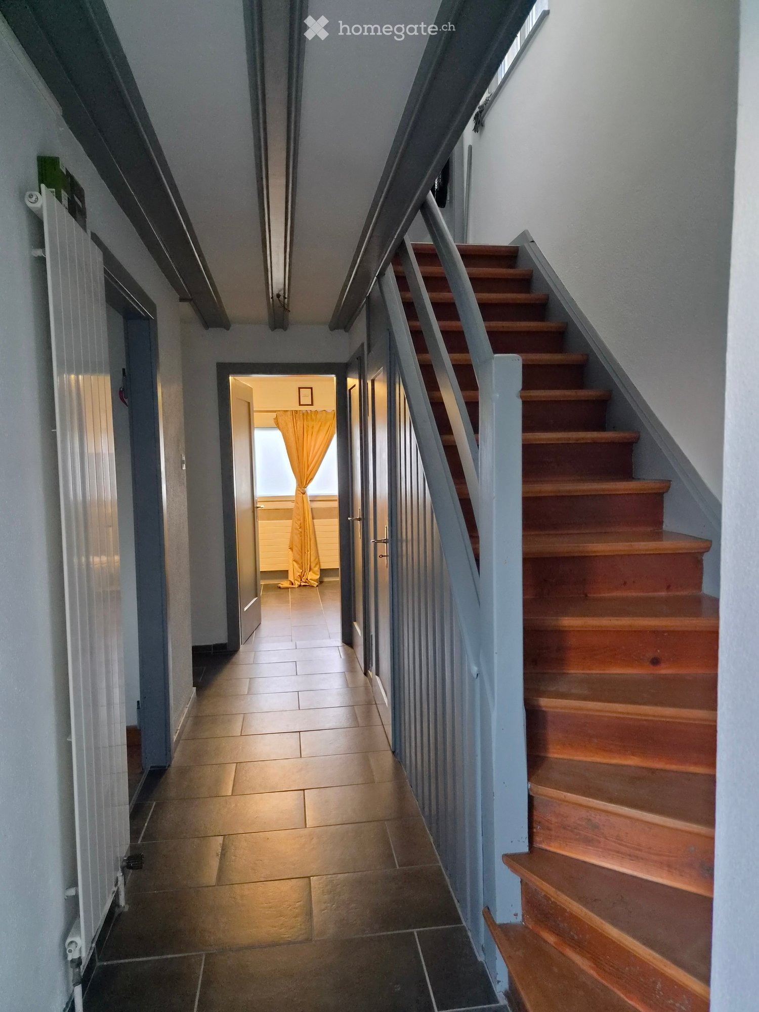A narrow hallway with tiled flooring, a wooden staircase with a metal railing, and an open doorway leading to another room with a yellow curtain.