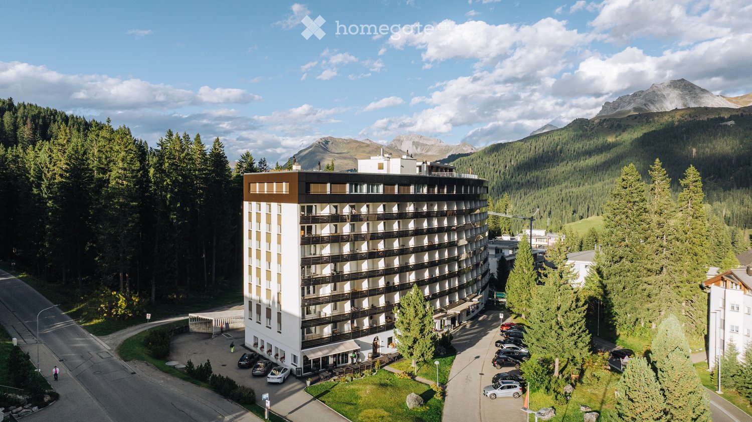 A modern multi-story hotel building with balconies, surrounded by trees and mountains, with a parking area and a few cars visible.