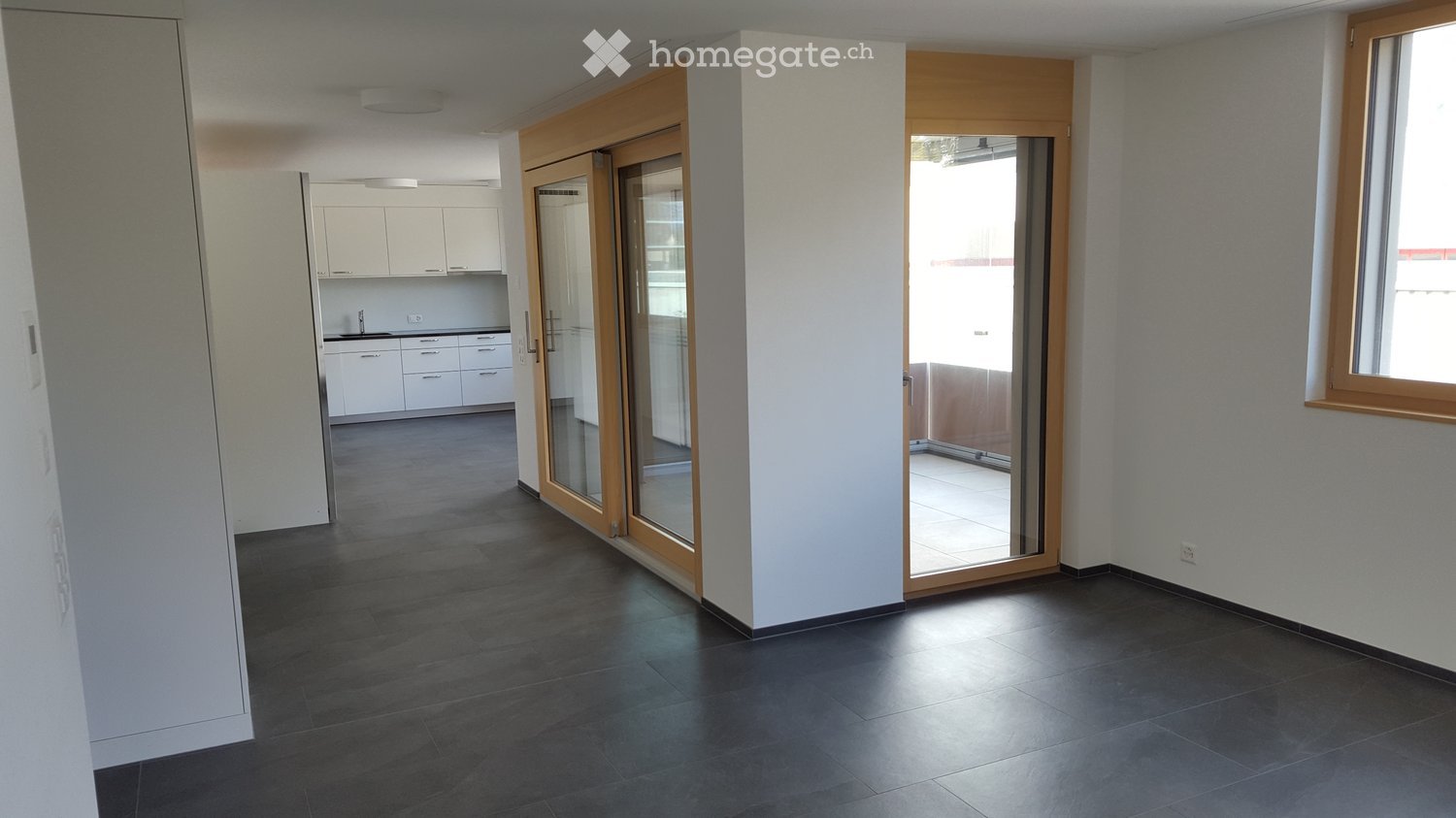 Empty hallway with dark tiled floor, white walls, and glass sliding doors leading to a balcony. Kitchen area visible in the background with white cabinets and countertops.