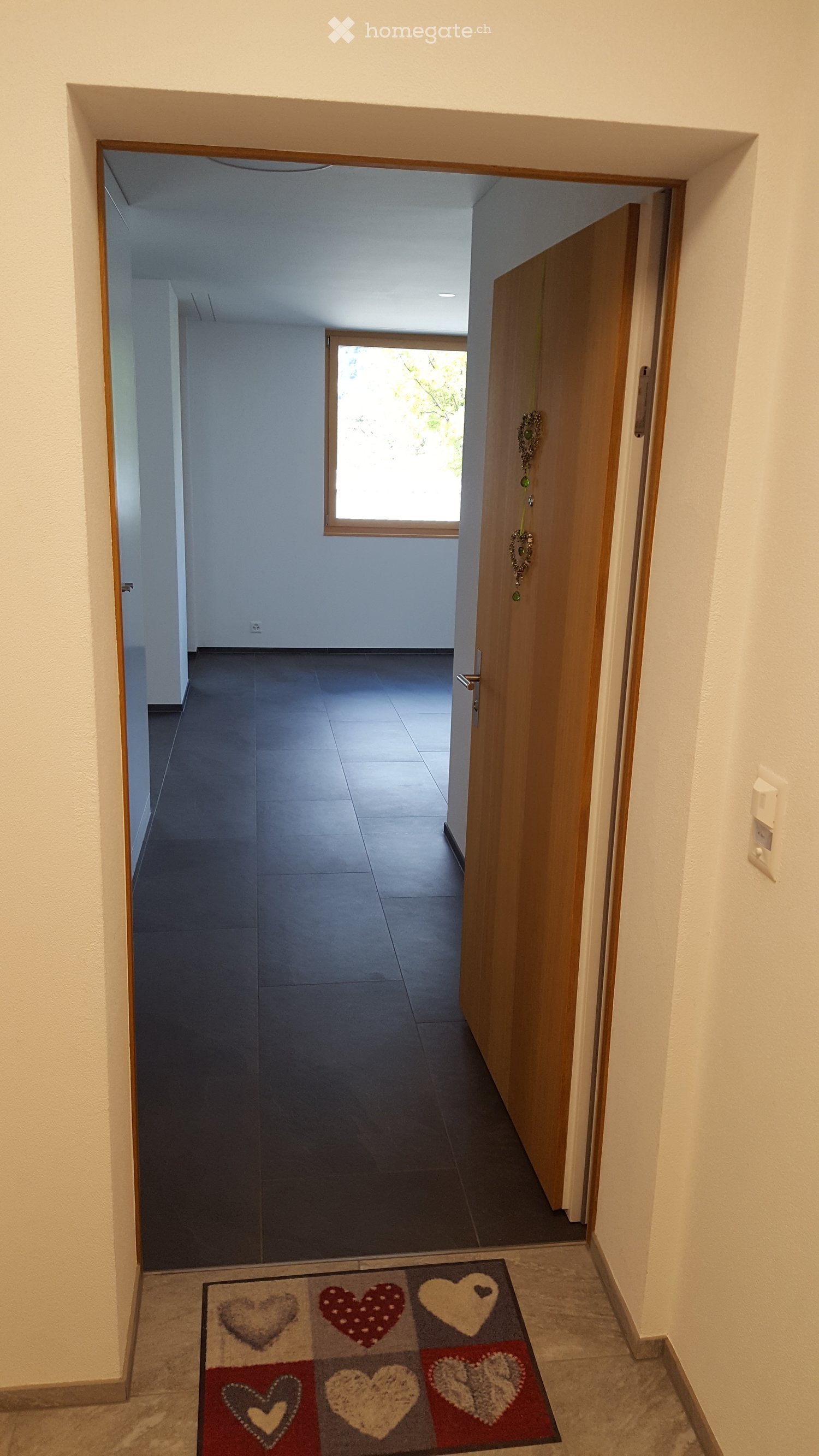 Empty hallway with tiled floor, light-colored walls, and a decorative rug featuring heart patterns near the entrance.