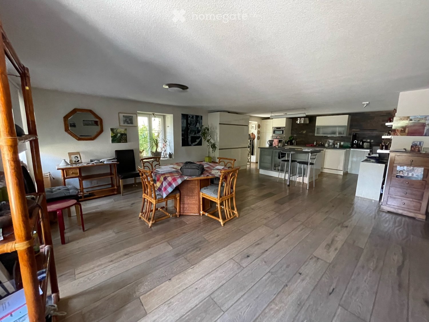The image shows a well-lit, open-plan living and dining area with wooden flooring. The dining area features a round wooden table with four matching chairs, a small wooden cabinet, and a red stool. The living area includes a black armchair, a small wooden