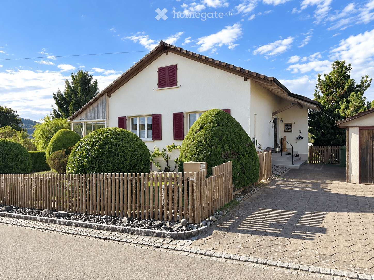 Single-story house with white exterior, red shutters, and a wooden fence. The house has multiple windows and a small porch with steps leading up to the entrance.