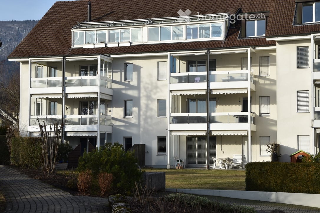 Modern multi-story apartment building with white exterior, multiple balconies, and a rooftop terrace. The building features large windows and a well-maintained garden area in front.
