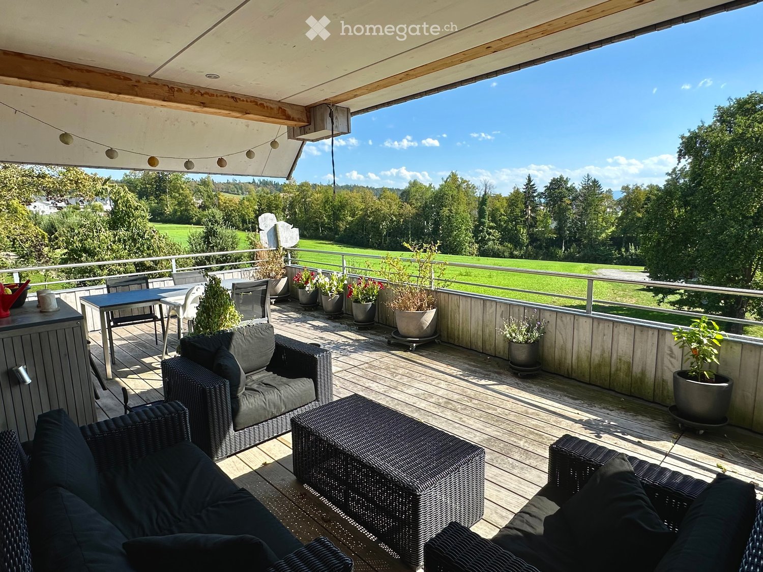 Wooden deck with outdoor furniture including a sofa, chairs, a table, and several potted plants, overlooking a green field and trees.
