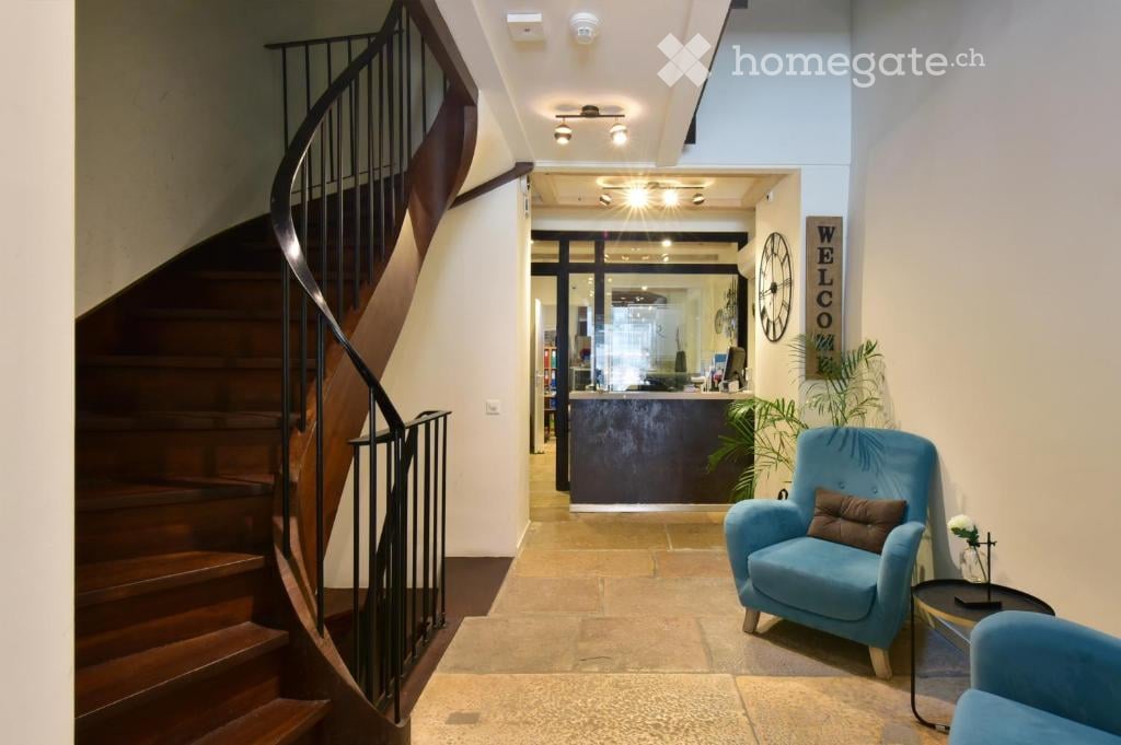 Spacious hallway with a wooden staircase, reception area, and seating area featuring a blue armchair and small table.
