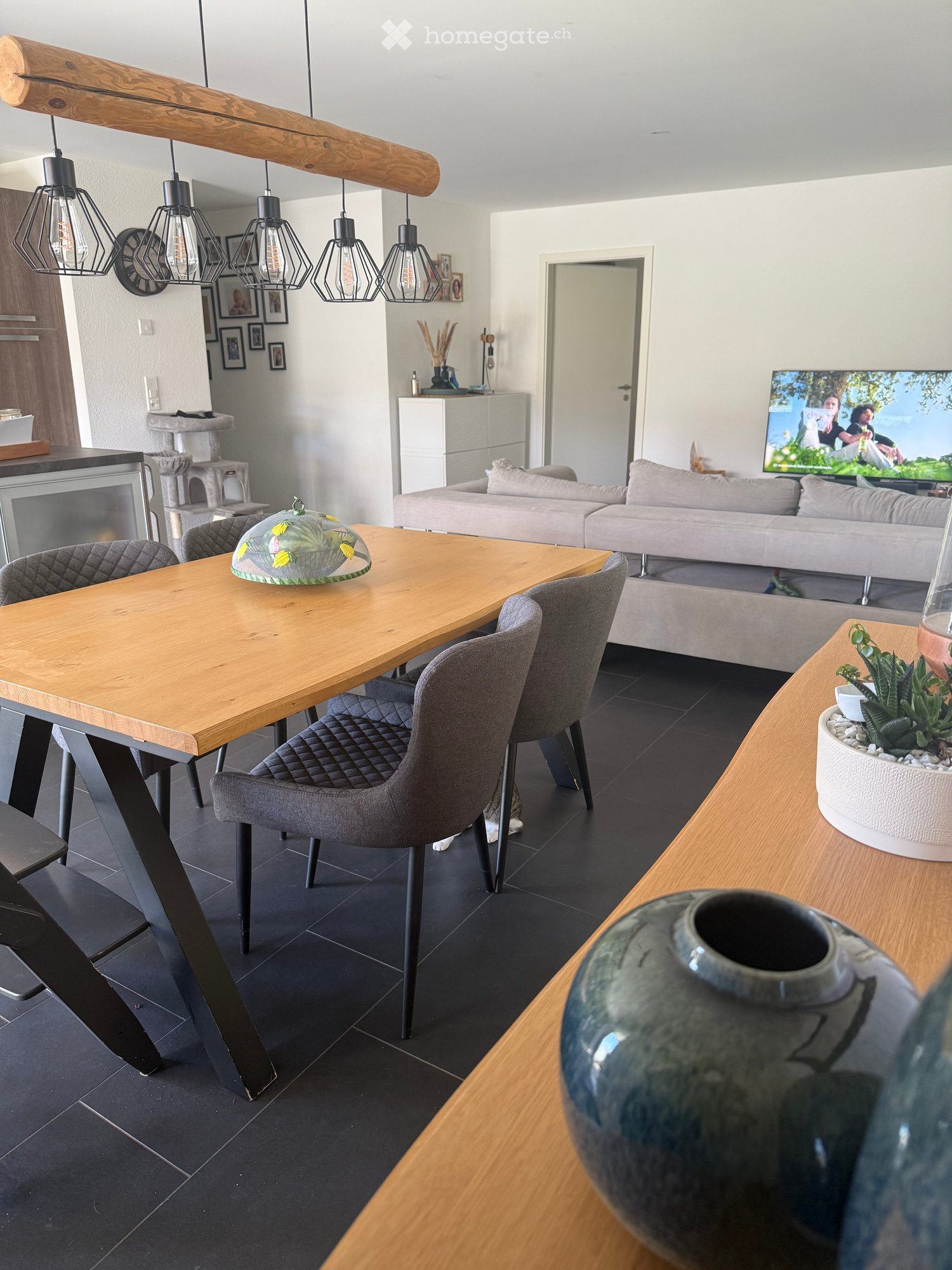 Modern living and dining area with a grey sofa, wooden dining table, grey chairs, television, pendant lights, and decorative vases.