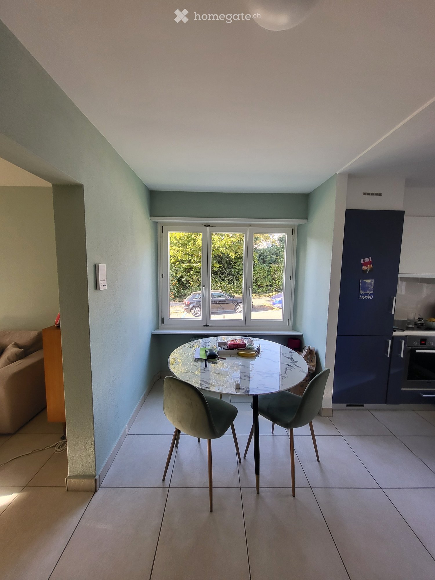 A modern, well-lit dining area with a round glass table, two green chairs, a sofa, and a kitchen area with a refrigerator and oven. Large windows provide natural light and a view of the outdoors.