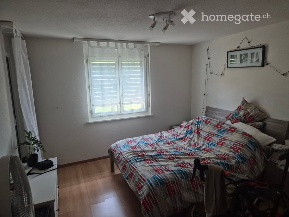 Bedroom with wooden flooring, a bed with a colorful quilt, a nightstand, a fan, a small plant, and a picture frame on the wall.