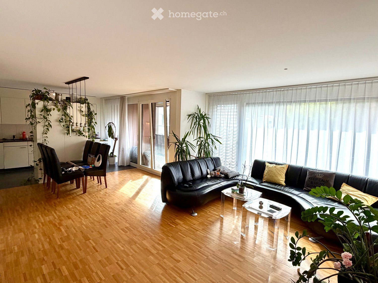 Modern living room with hardwood flooring, large windows, and a balcony. Features a black leather sofa, glass coffee tables, and several chairs.