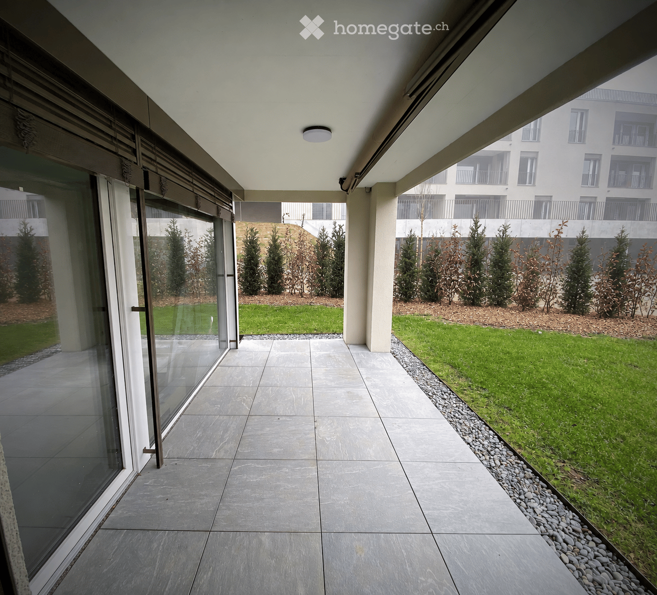 Modern covered balcony with tiled flooring, large glass sliding doors, and a view of a landscaped garden with small trees and shrubs.