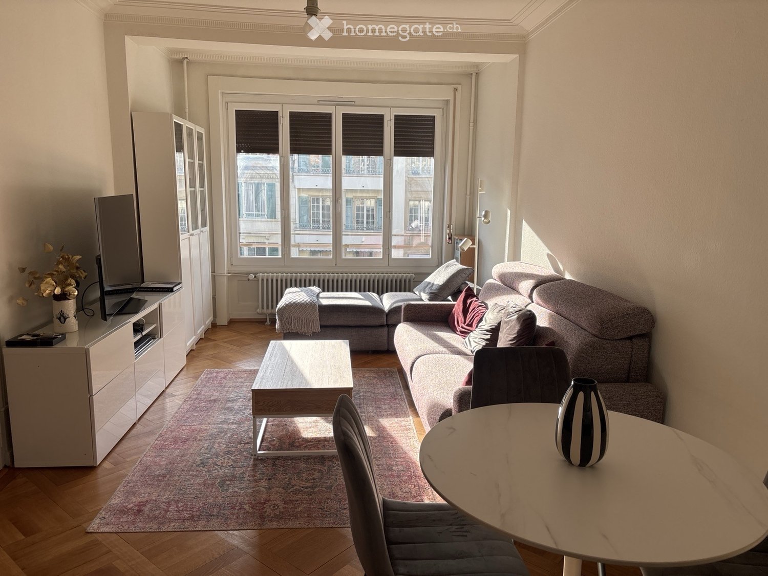 Bright living room with wooden flooring, large windows, grey sectional sofa, coffee table, dining table with chairs, and a TV cabinet.