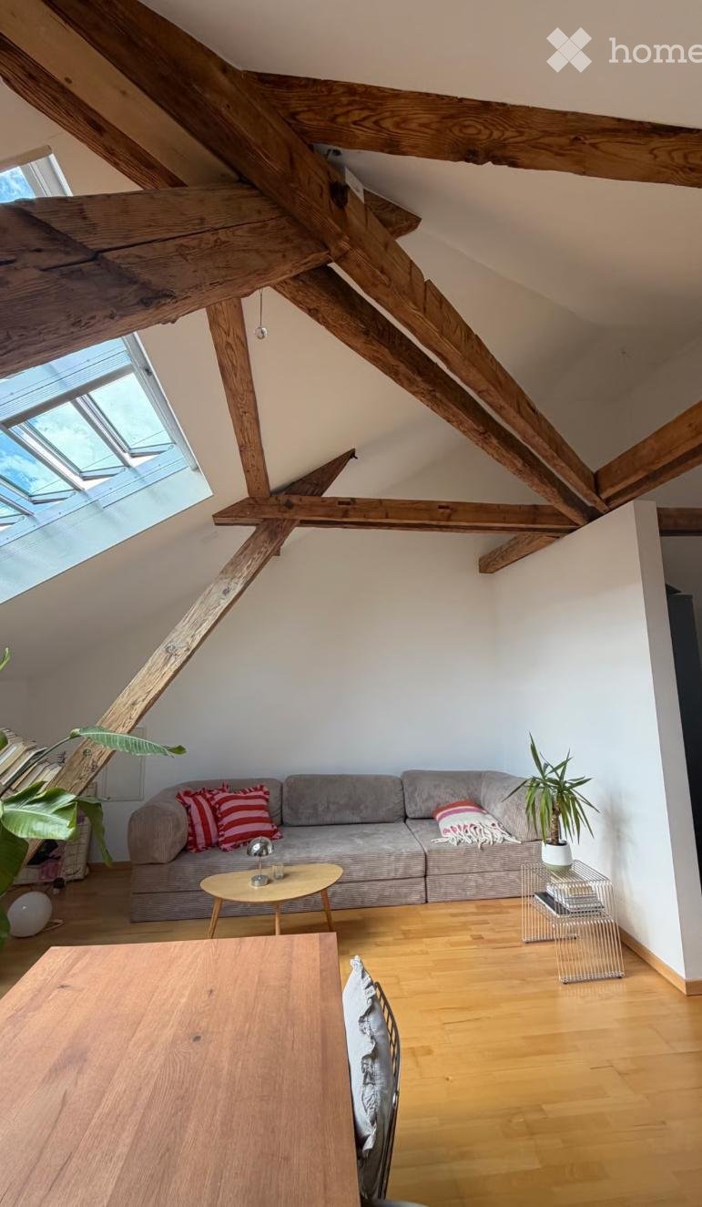 Bright living room with exposed wooden beams, grey sofa with red pillows, small coffee table, wooden dining table, and potted plants.