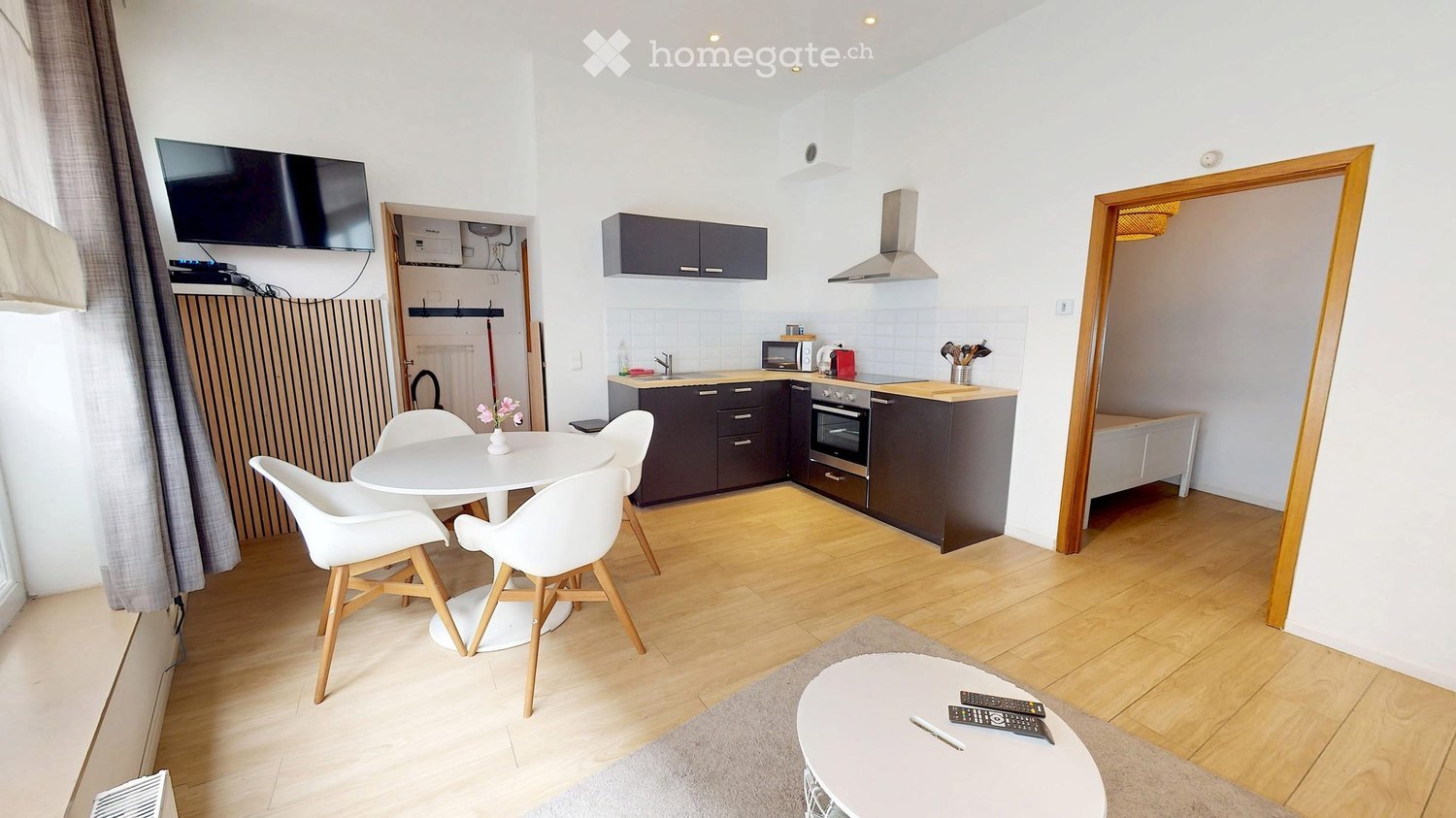 Modern, open-plan living and dining area with a white dining table, four chairs, a small round coffee table, a television, and a compact kitchen featuring dark cabinets, a stainless steel range hood, and built-in appliances.